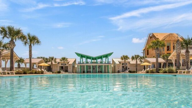 A resort pool area with clear blue water, palm trees, and a building with a green roof. - Game Room, Resort Pools, Golf Cart Boardwalk to the Beach (Port Aransas)