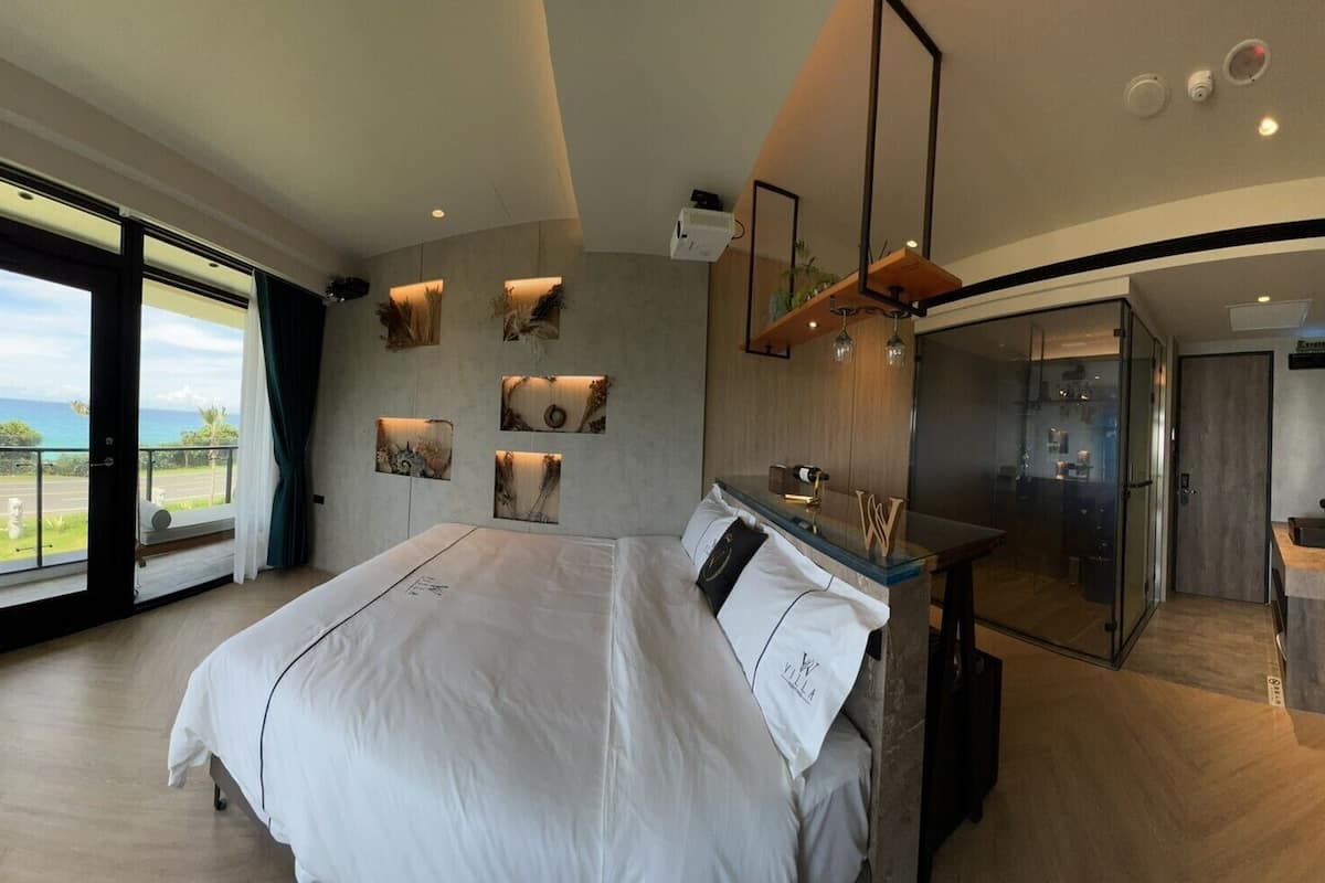 A modern hotel room with a large bed, a glass shower, and a balcony with a view.