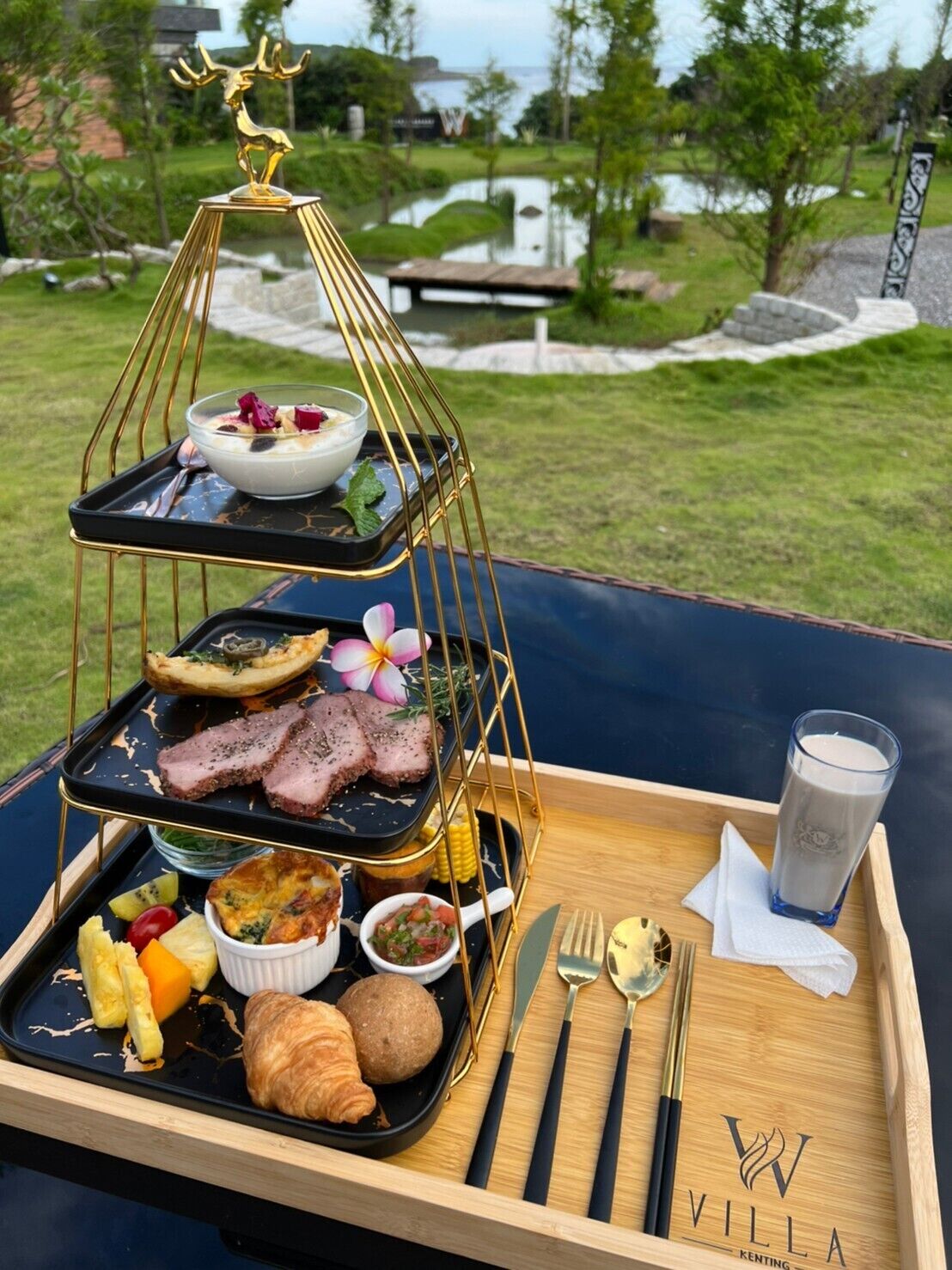 A three-tiered serving tray with food and drinks, a glass of milk, and cutlery on a wooden tray with a logo.