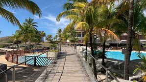 A wooden boardwalk leading to a pool area with palm trees and a building in the background.