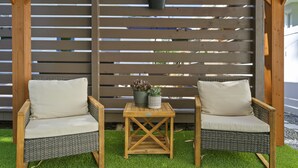 A wooden pergola with horizontal slats, two wicker chairs with cushions, and a small table with potted plants. - Caffeinated Bungalito 75in TV- Walkable to Armature Works (Tampa)