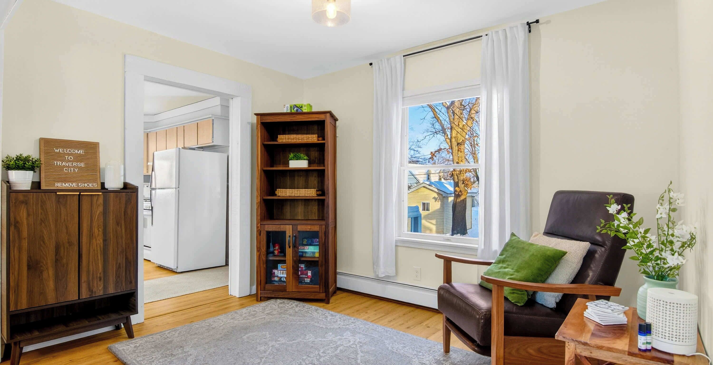 A living room with a wooden cabinet, a bookshelf, a leather chair, a window with curtains, and a small table with a plant.