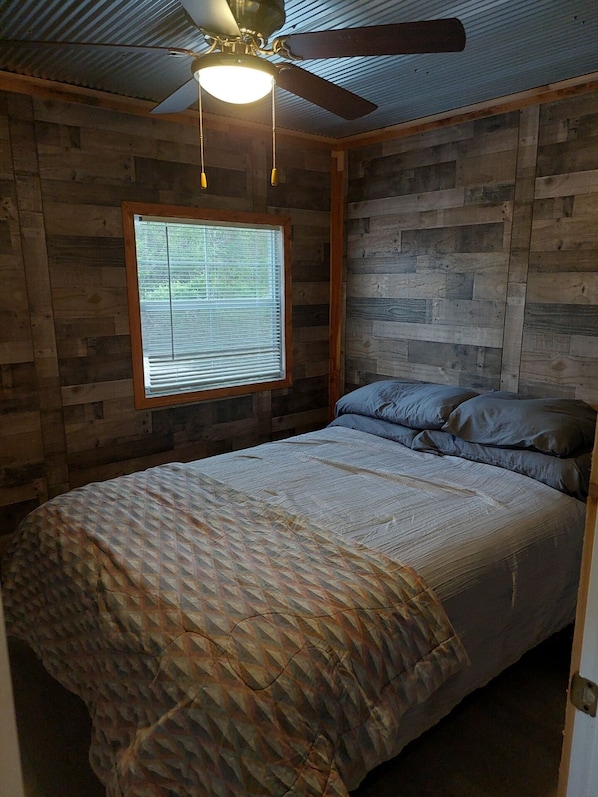 A bedroom with a wooden wall, a ceiling fan, a window with blinds, and a bed with a patterned blanket.