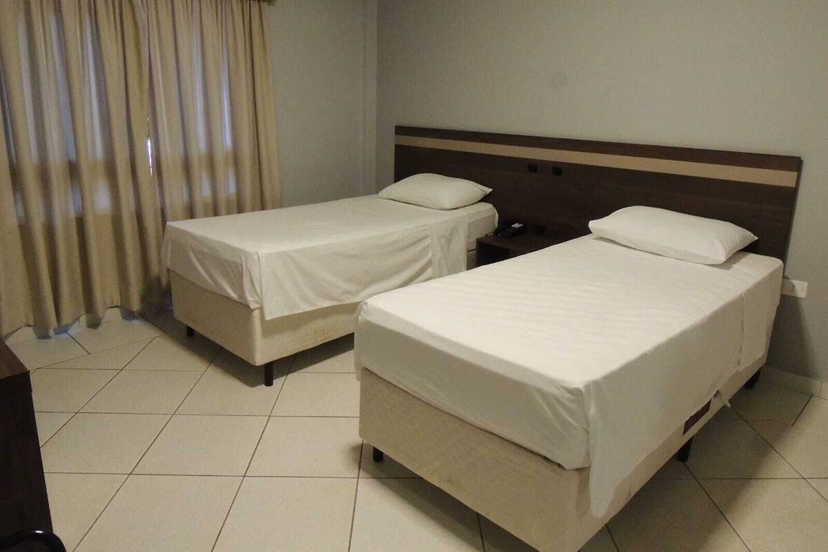 Una habitación de hotel con dos camas individuales, una mesita de noche y un ventanal con cortinas.