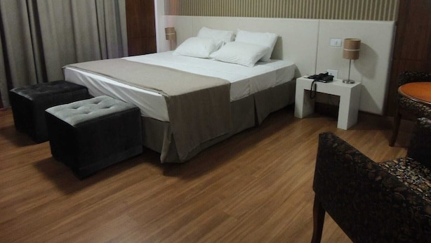 A hotel room with a bed, two ottomans, a nightstand with a lamp, and a chair. - Vilhar Palace Hotel (Ivaipora)