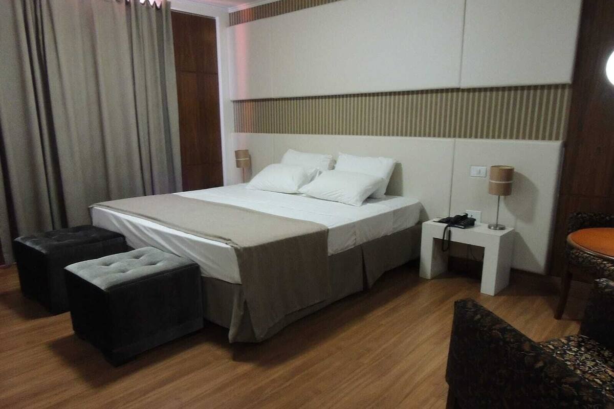 Una habitación de hotel con una cama, dos otomanes, una mesita de noche con lámpara y una silla.