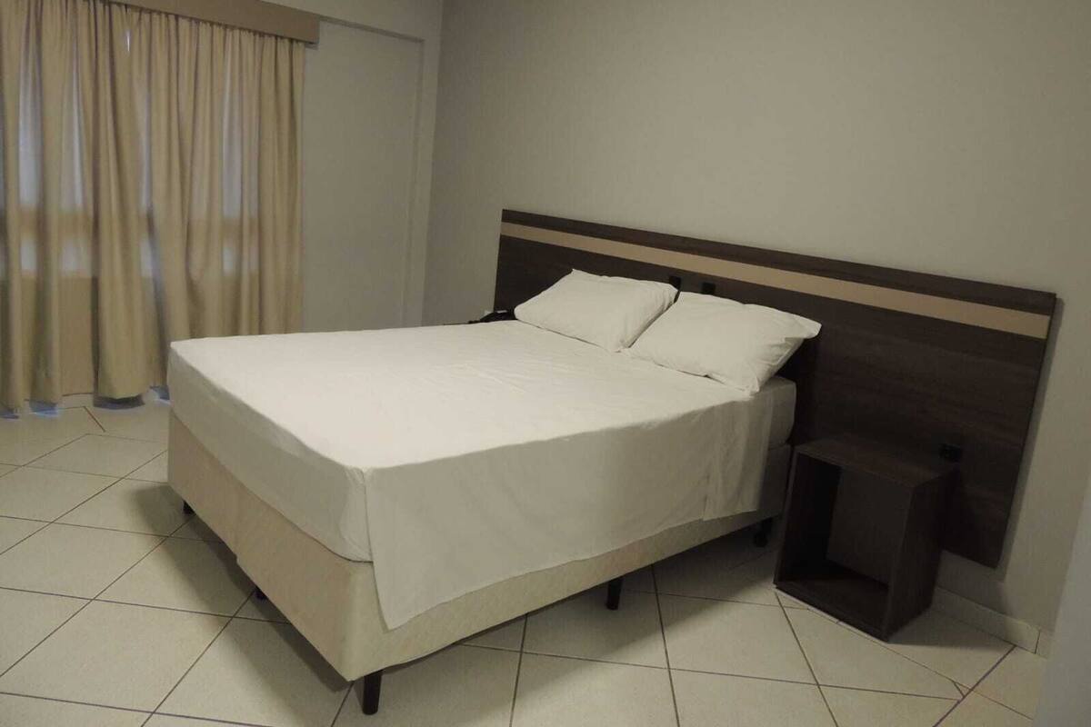 Una cama bien tendida con cabecera de madera en una habitación de hotel con suelo de baldosas y una ventana con cortinas.