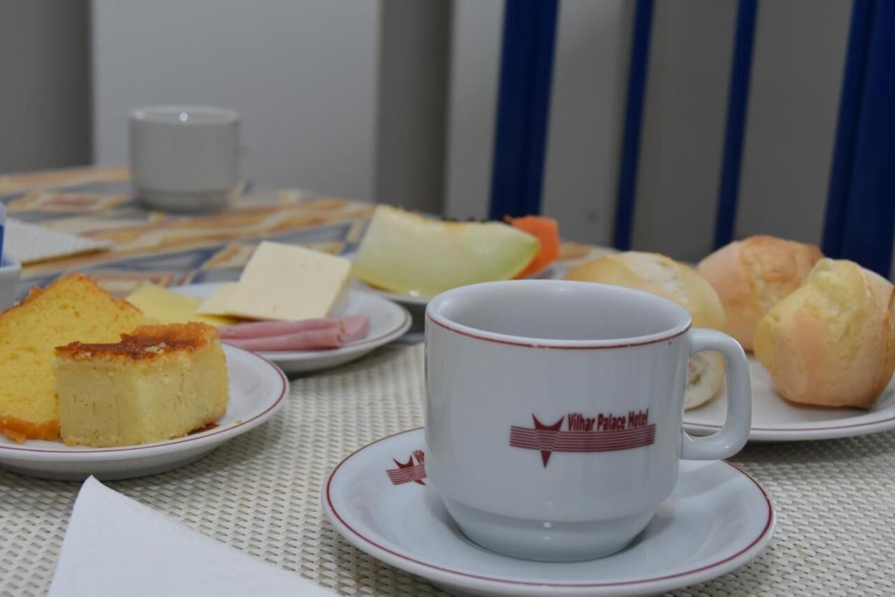 A table with a white cup and saucer, a slice of cake, a croissant, and a small plate with a piece of ham.