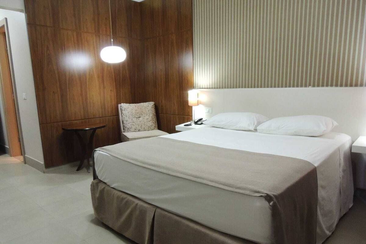 Habitación de hotel con una cama grande, una mesita de noche con lámpara, una silla y un panel de pared de madera.