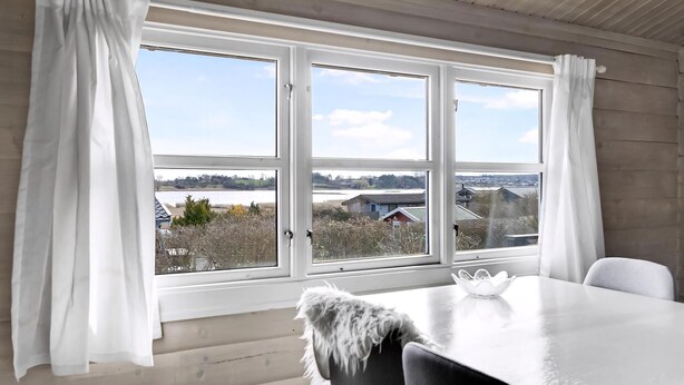 Una habitación con un ventanal grande, cortinas blancas, una mesa blanca y un edredón mullido, con vistas a un lago y edificios.