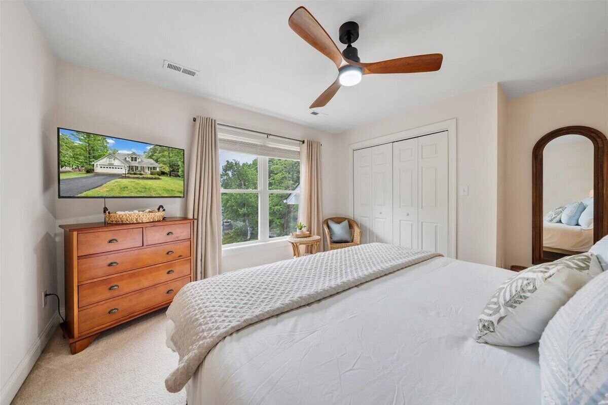 A bedroom with a bed, a wooden dresser, a television, a ceiling fan, a window with curtains, and a door.