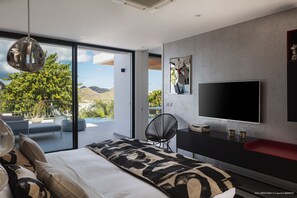 A modern bedroom with a large bed, a TV, a chair, and a view of a balcony with plants and a mountain. - Villa Ventura | Ocean View - Located in  Magnificent Lurin with Private Pool (Lurin)