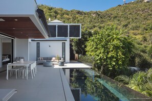 A modern villa with a glass infinity pool, outdoor seating, and a view of a lush hillside.