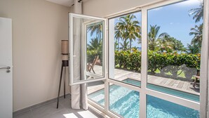 A room with a large sliding glass door leading to a pool area, a standing lamp, and a view of palm trees and a wooden deck.
