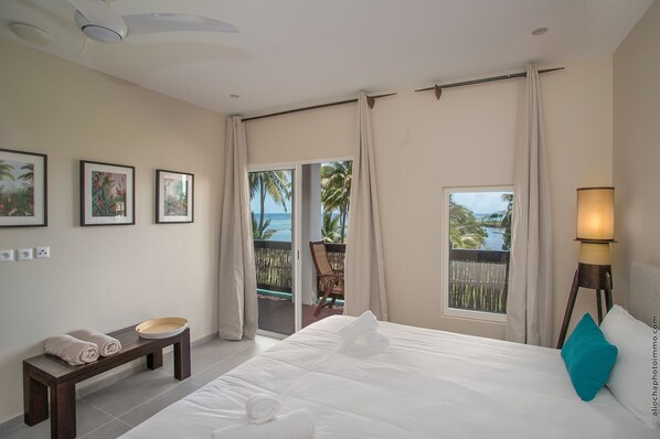 A hotel room with a bed, a bench with towels, a lamp, and a view of the ocean through the balcony doors.