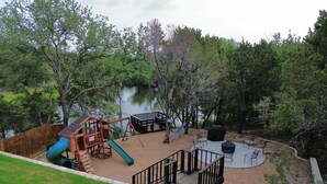 A playground with a slide and swings, a covered picnic table, and a wooden deck overlooking a lake. - Scenic Retreat W/ Playground & Grilling (Granbury)