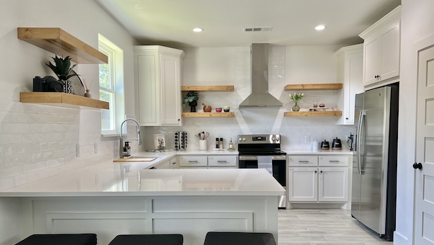 A modern kitchen with white cabinetry, a large island with bar stools, stainless steel appliances, and wooden shelves. - Scenic Retreat W/ Playground & Grilling (Granbury)