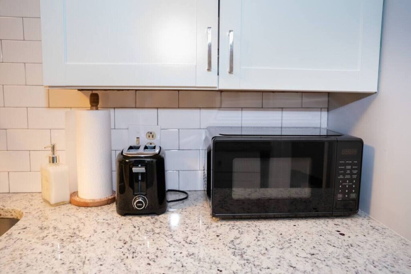 Une cuisine avec des meubles blancs, un grille-pain noir, un four à micro-ondes et un distributeur blanc posé sur un plan de travail en granit.