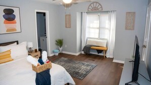 Une chambre d’hôtel équipée d’un lit, d’une télévision, d’un ventilateur de plafond, d’une fenêtre avec stores, d’un petit banc et d’un téléviseur fixé au mur.