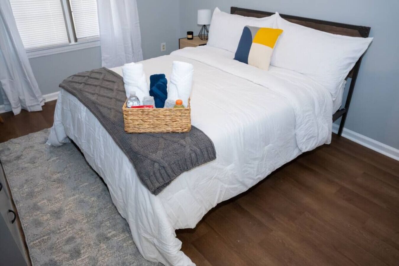 A neatly made bed with a grey blanket, a wicker basket with toiletries, and a bedside table with a lamp.