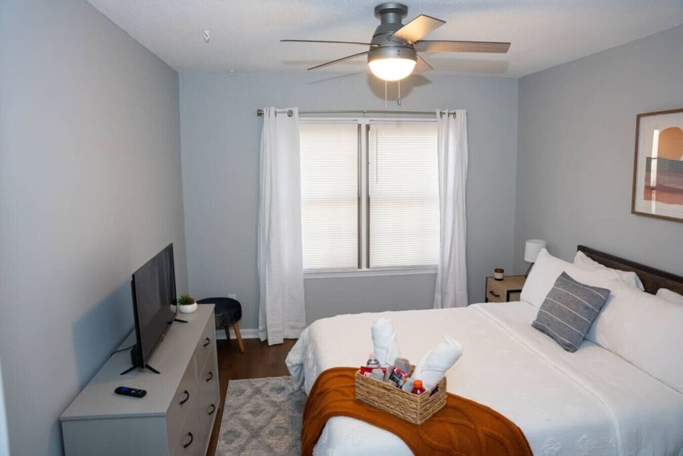 A bedroom with a bed, a ceiling fan, a TV, a desk, and a window with curtains.