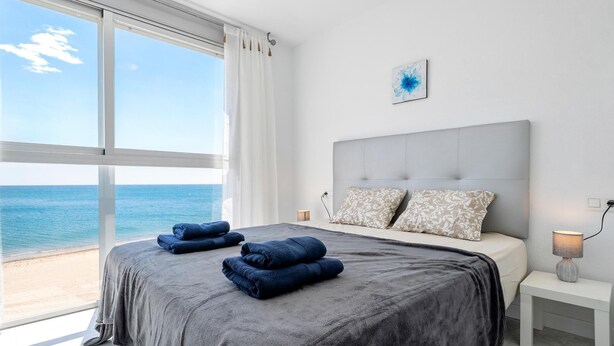 Ein Schlafzimmer mit großem Fenster mit Meerblick, einem Bett mit grauer Bettwäsche, zwei Kissen und einem Nachttisch mit Lampe.