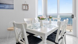 Ein Essbereich mit einem weißen Tisch und Stühlen, einer Vase mit Blumen und Blick auf das Meer durch große Fenster.