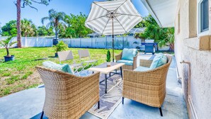 Une terrasse extérieure aménagée avec des meubles en osier, un parasol à rayures et une vue sur un jardin avec une clôture et des arbres.