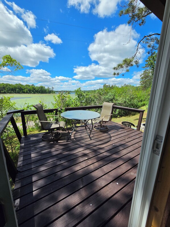 Une terrasse en bois avec une table ronde et des chaises, offrant une vue sur un paysage verdoyant et une étendue d’eau sous un ciel bleu parsemé de nuages.
