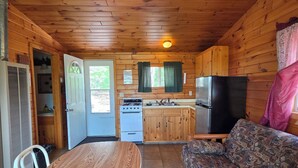 A cabin interior with a dining table, chairs, a sofa, a kitchen with a stove and refrigerator, and a ceiling fan.