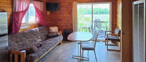 A cabin interior with a wooden ceiling, a sofa, a table with chairs, a television, and a window with curtains.