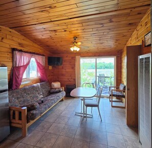 A cabin interior with a wooden ceiling, a sofa, a table with chairs, a television, and a window with curtains. - East Crooked Lake Cabin 3 (Nevis)