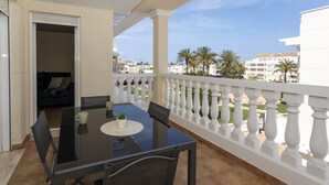 Uma varanda com mesa e cadeiras de vidro, vista para palmeiras e edifícios, e um guarda-corpo branco.