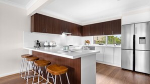 A modern kitchen with dark wood cabinets, a white island with stools, stainless steel appliances, and a view of the outdoors. - Meira Haus - Belle Escapes Jervis Bay (Sanctuary Point)
