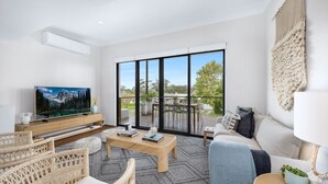 A modern living room with a large TV, comfortable seating, a wooden coffee table, and a balcony with outdoor furniture. - Meira Haus - Belle Escapes Jervis Bay (Sanctuary Point)