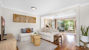 A modern living room with a white sofa, a coffee table, and a wooden side table. There is a large window with a view of greenery outside. - Meira Haus - Belle Escapes Jervis Bay (Sanctuary Point)