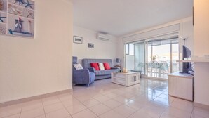 A modern living room with a grey sofa, a white coffee table, and a TV stand. There is a large window with a view of the outside.