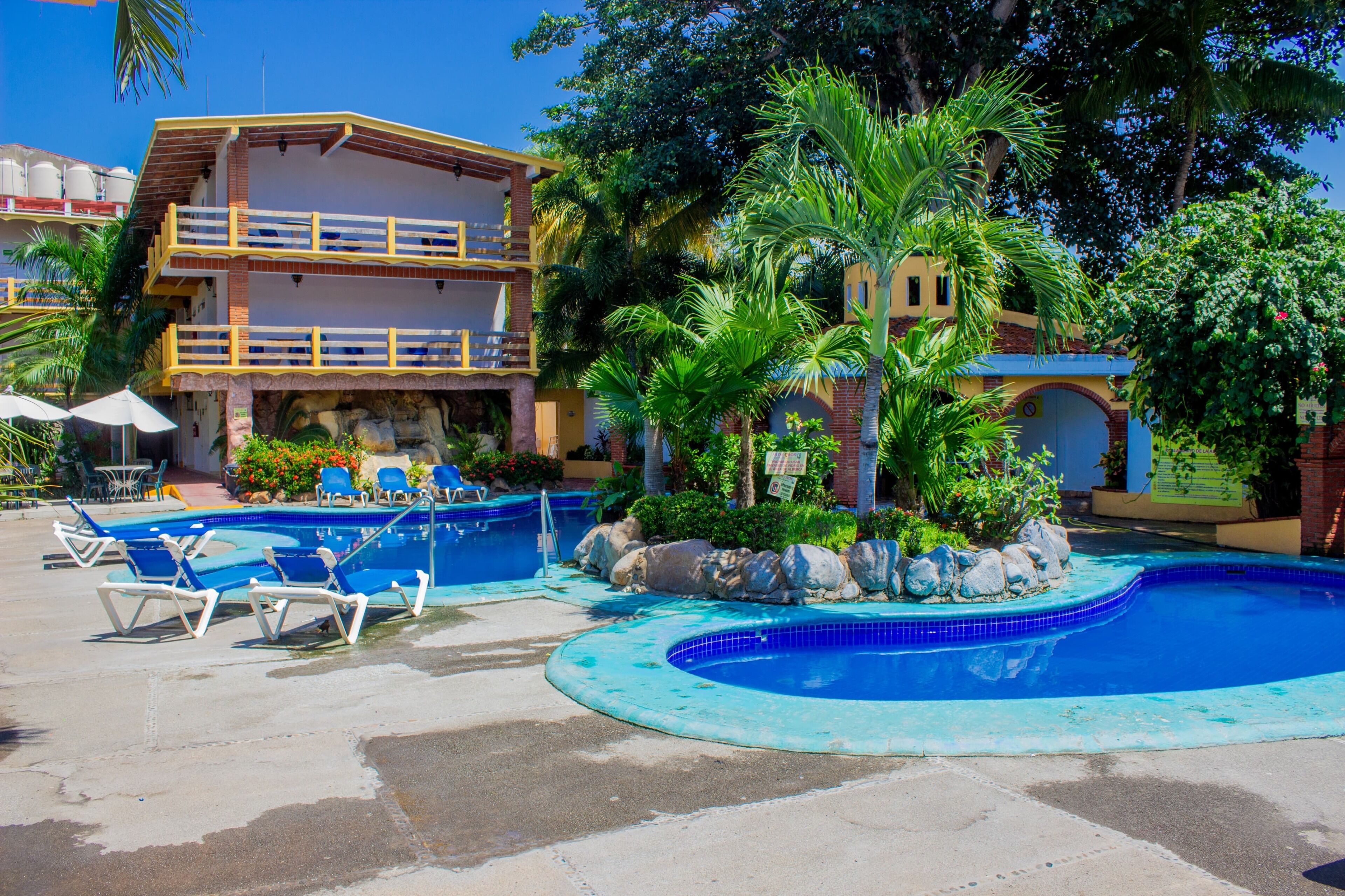 Un espace piscine avec des chaises longues, un bâtiment doté d’un balcon et une végétation tropicale.