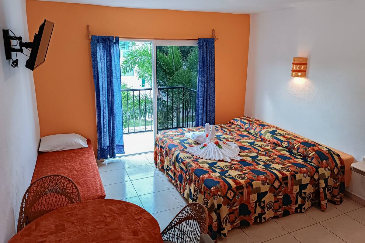 Une chambre d’hôtel avec un mur orange, un lit recouvert d’une courtepointe colorée, une petite table avec un coussin rouge, une chaise et un balcon avec des rideaux bleus.