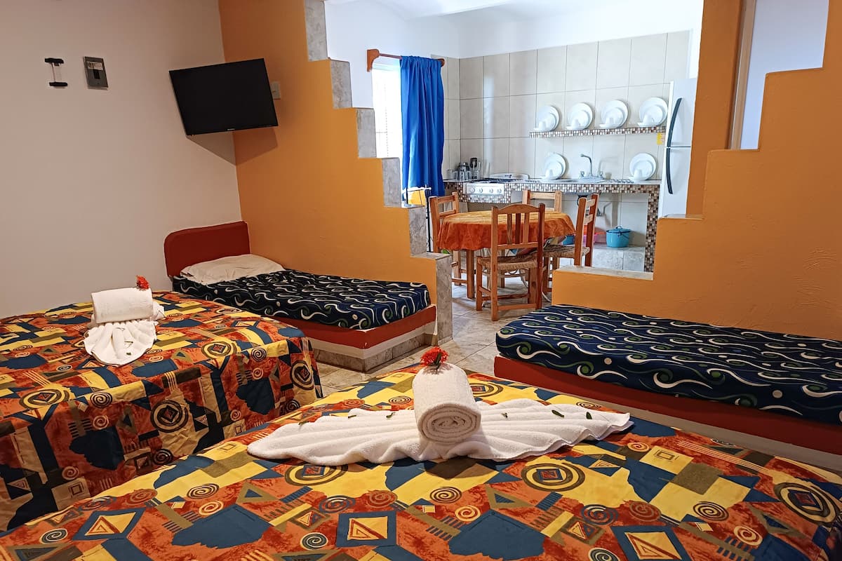 Une chambre d’hôtel avec deux lits, une télévision, un coin repas avec une table et des chaises, et une cuisine visible à travers une porte ouverte.
