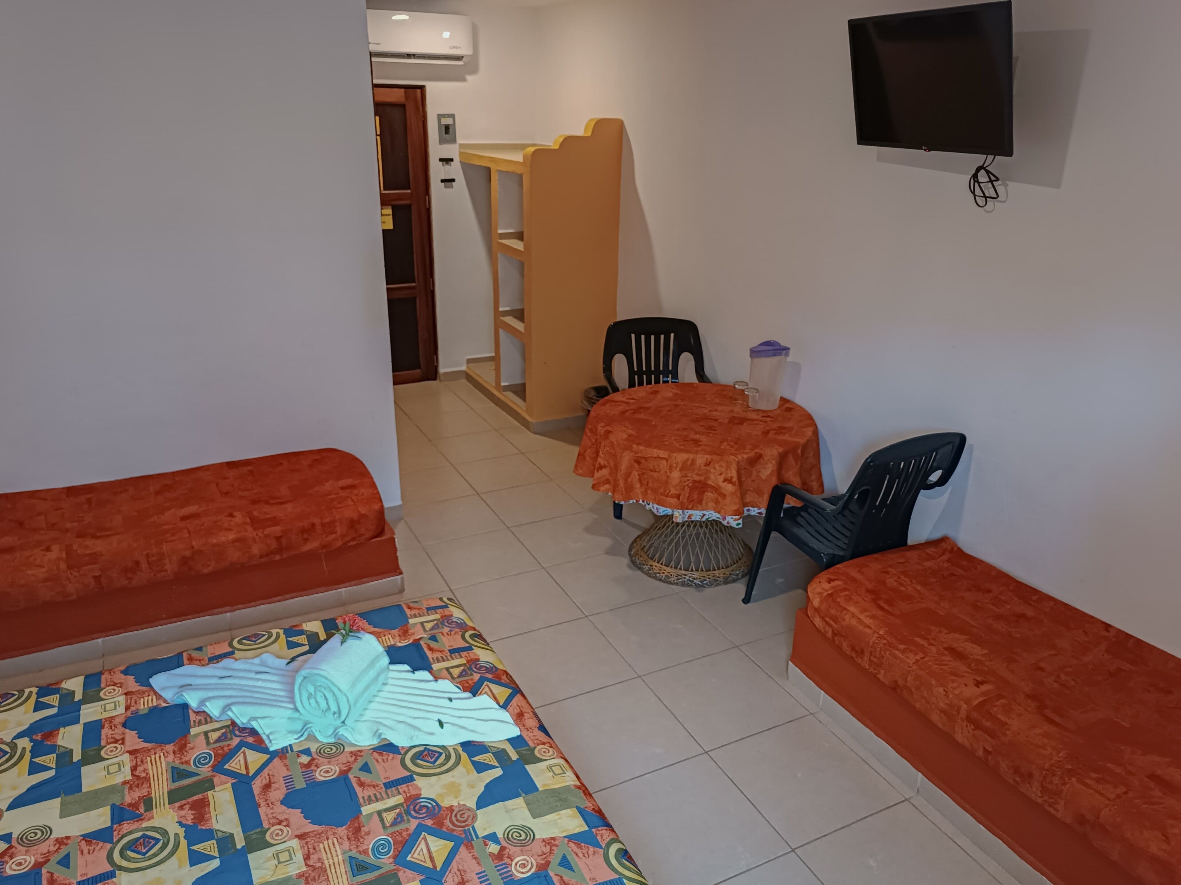 A room with two orange sofas, a small table with a red tablecloth, a TV mounted on the wall, and a patterned rug on the floor.