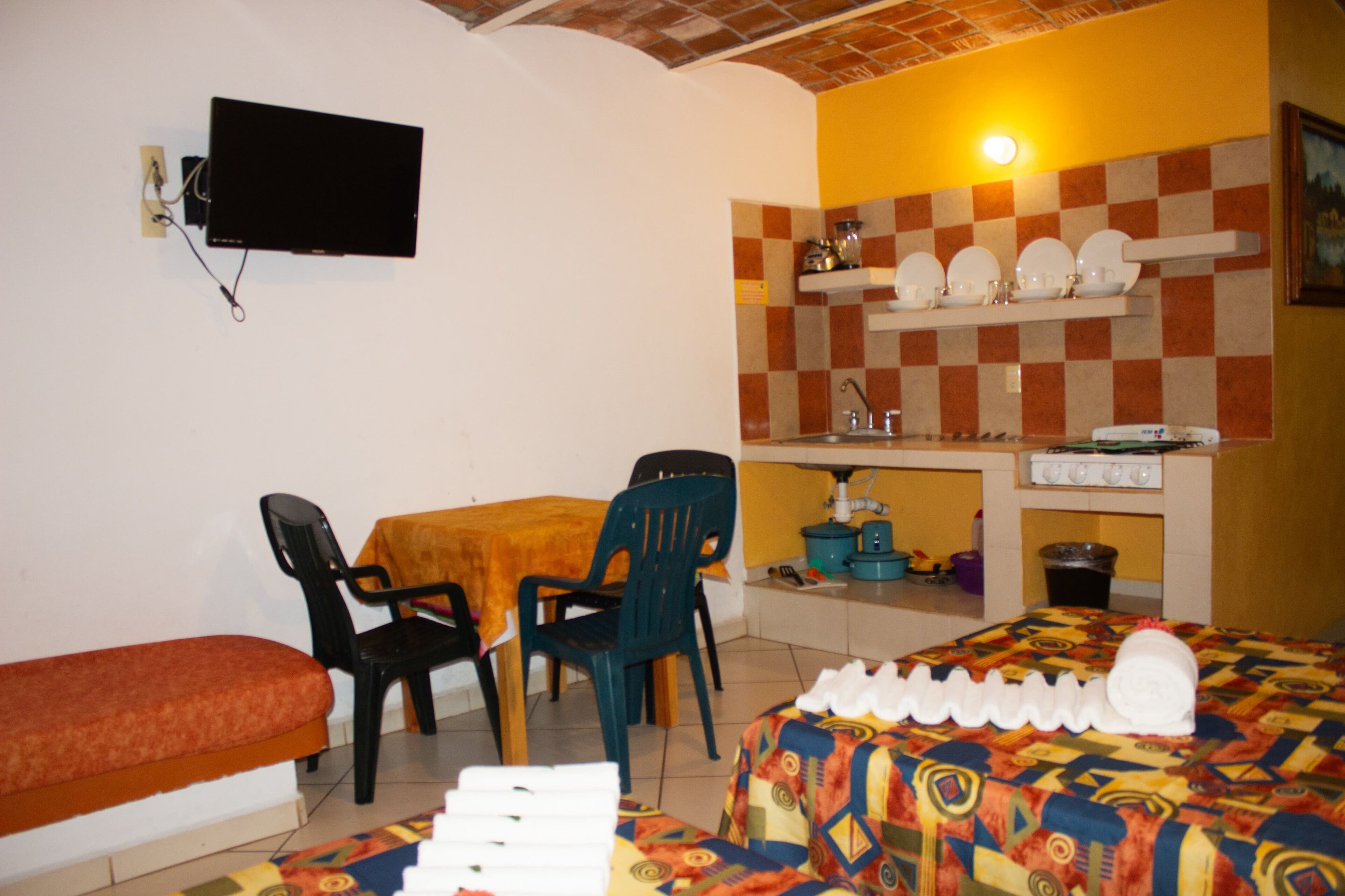 Ein kleines Zimmer mit einem Bett, einem Tisch mit Stühlen, einem an der Wand befestigten Fernseher und einer Küchenzeile mit Regalen über der Arbeitsfläche.