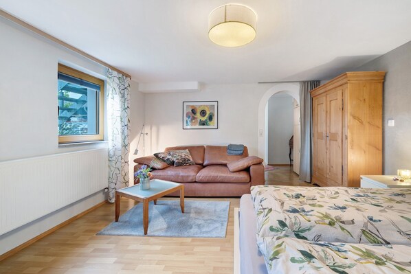 A room with a brown leather sofa, a wooden table, a bed with floral bedding, a large window with curtains, a wooden wardrobe, and a painting on the wall.