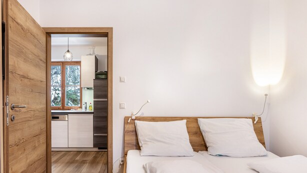 A bedroom with a wooden door, a bed with white bedding, a wooden headboard, and a view of a kitchen and window. - Holiday Apartment "Goodlife Top 1" with Mountain View & Wi-Fi (Saalbach)
