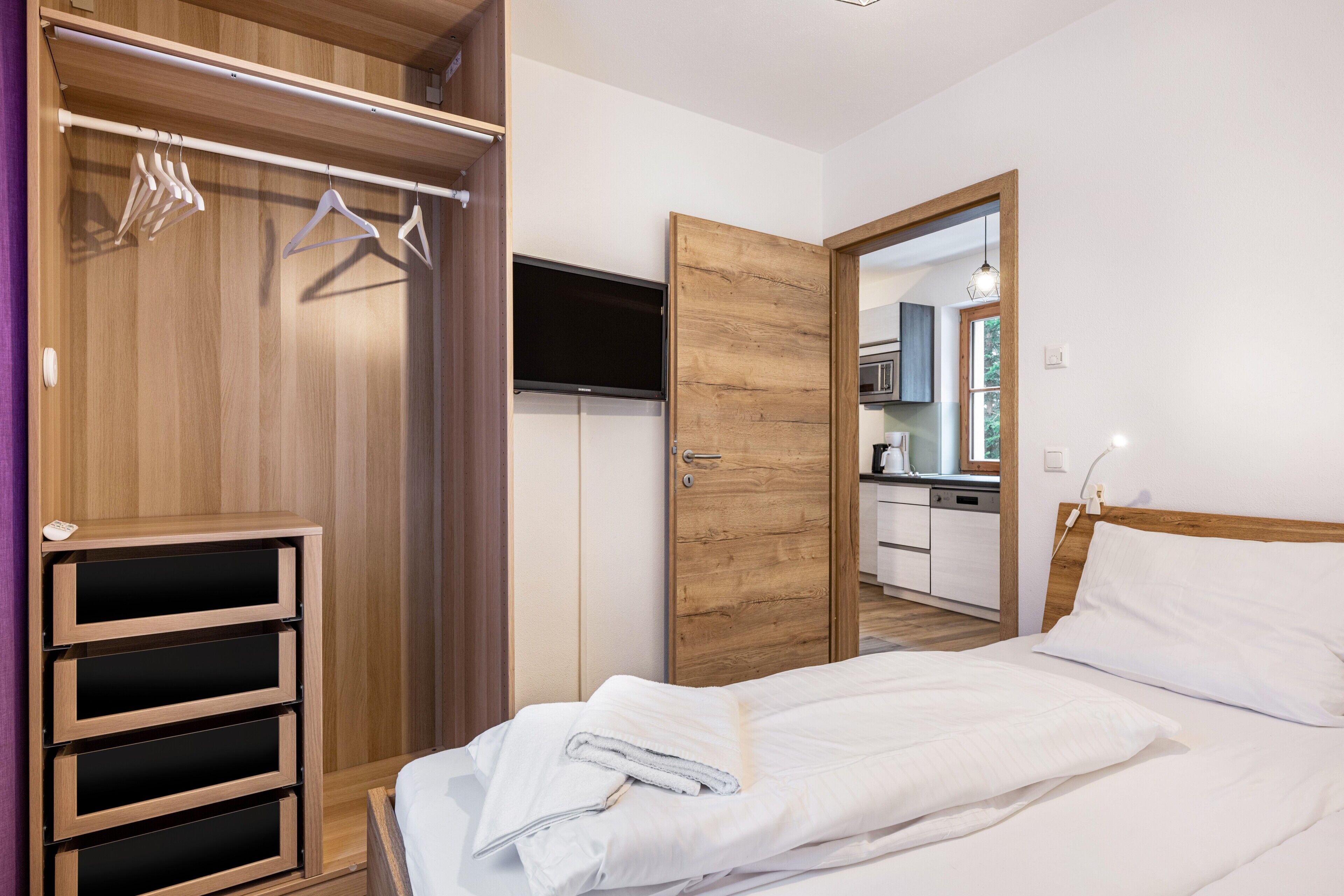 Un dormitorio con armario de madera, televisión, cama con ropa blanca y vista a la cocina.