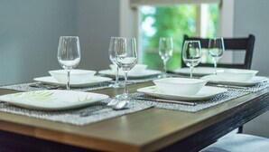 Mesa de jantar posta com pratos, tigelas e copos brancos, com vista para o verde pela janela.