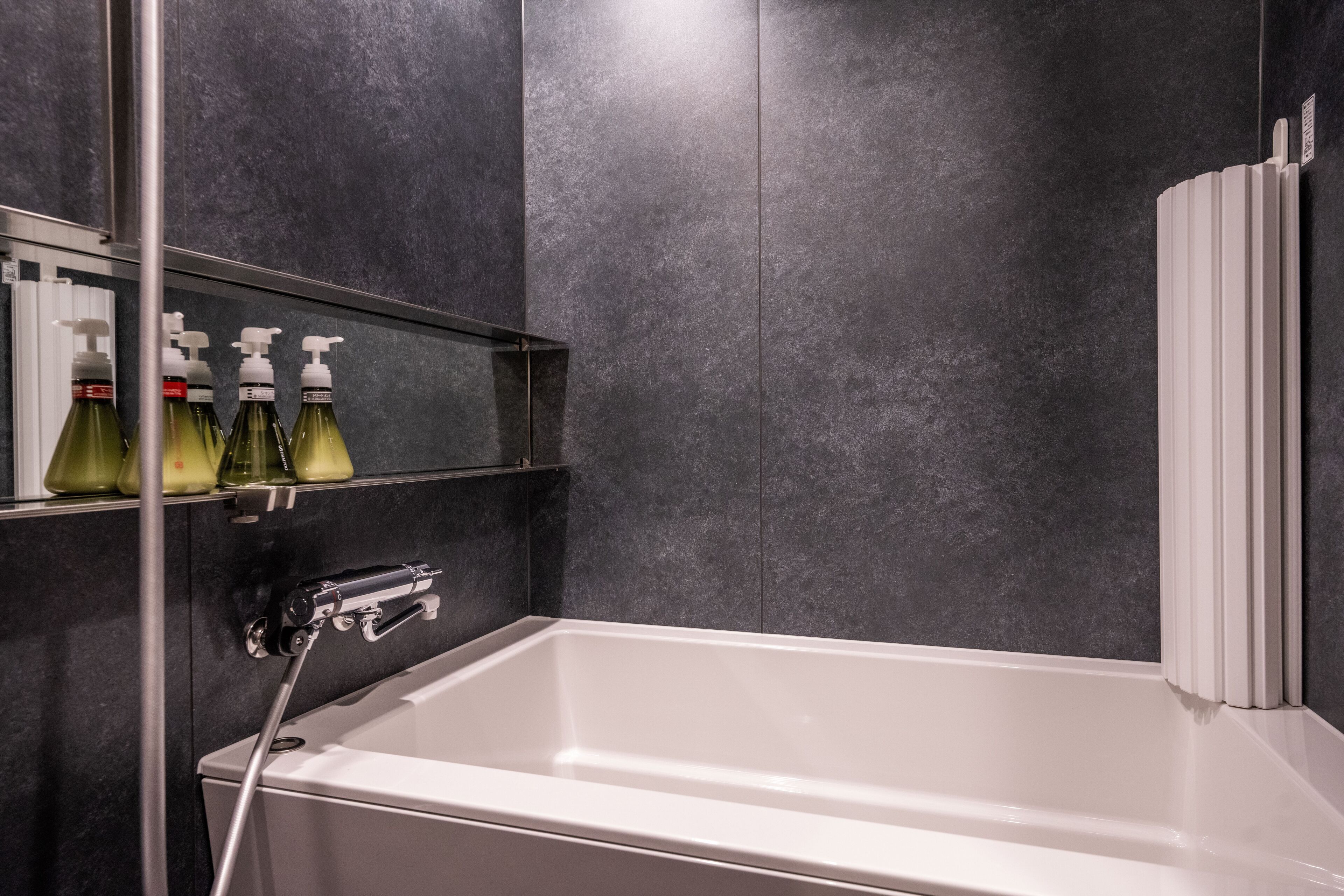 A modern bathroom with a white bathtub, dark tiled walls, and a shelf with bottles.