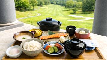 A tray with various Japanese dishes, including rice, salmon, and vegetables, set against a scenic view of a green landscape.