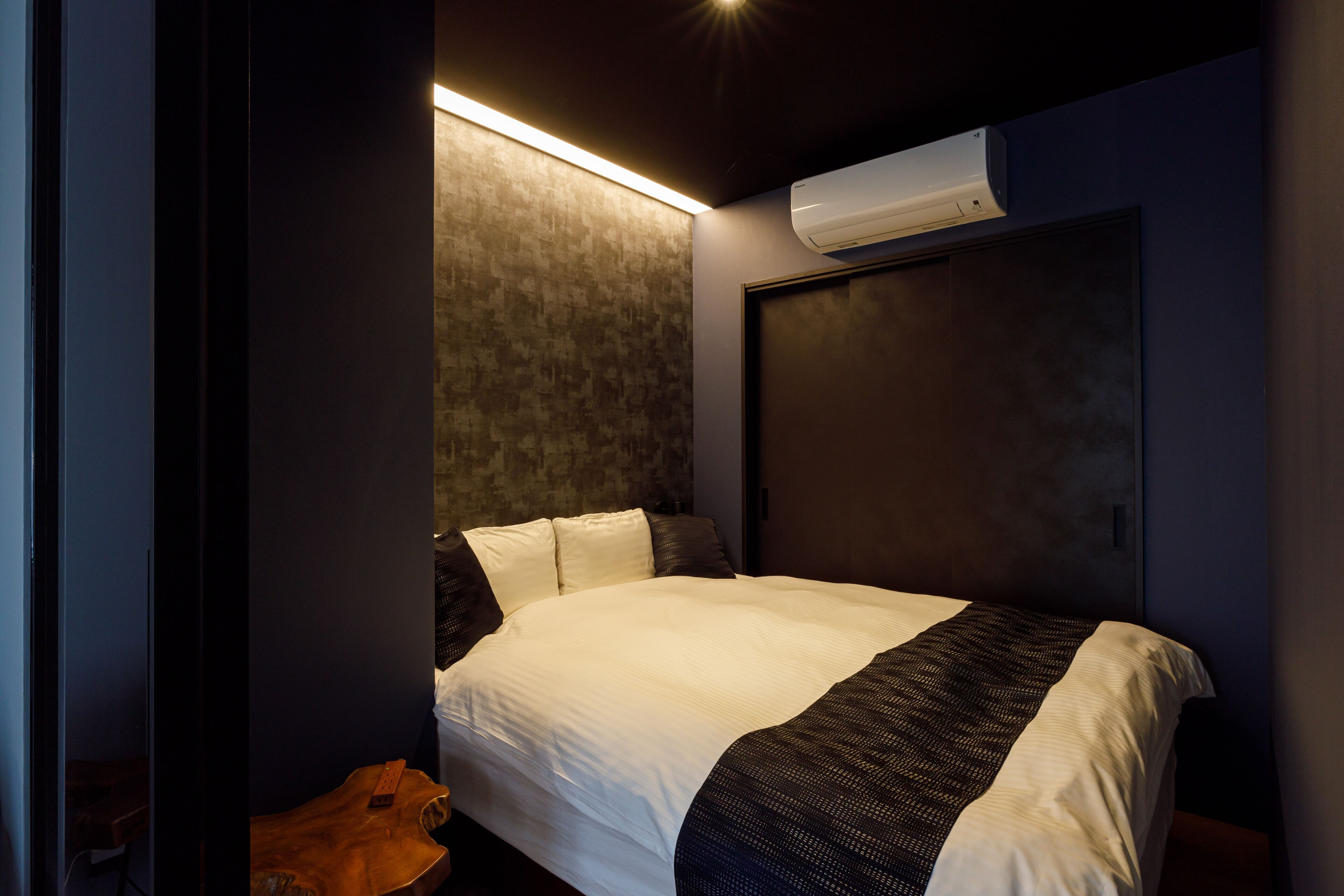 A modern hotel room with a large bed, a wooden side table, a wall-mounted air conditioner, and a textured wall.