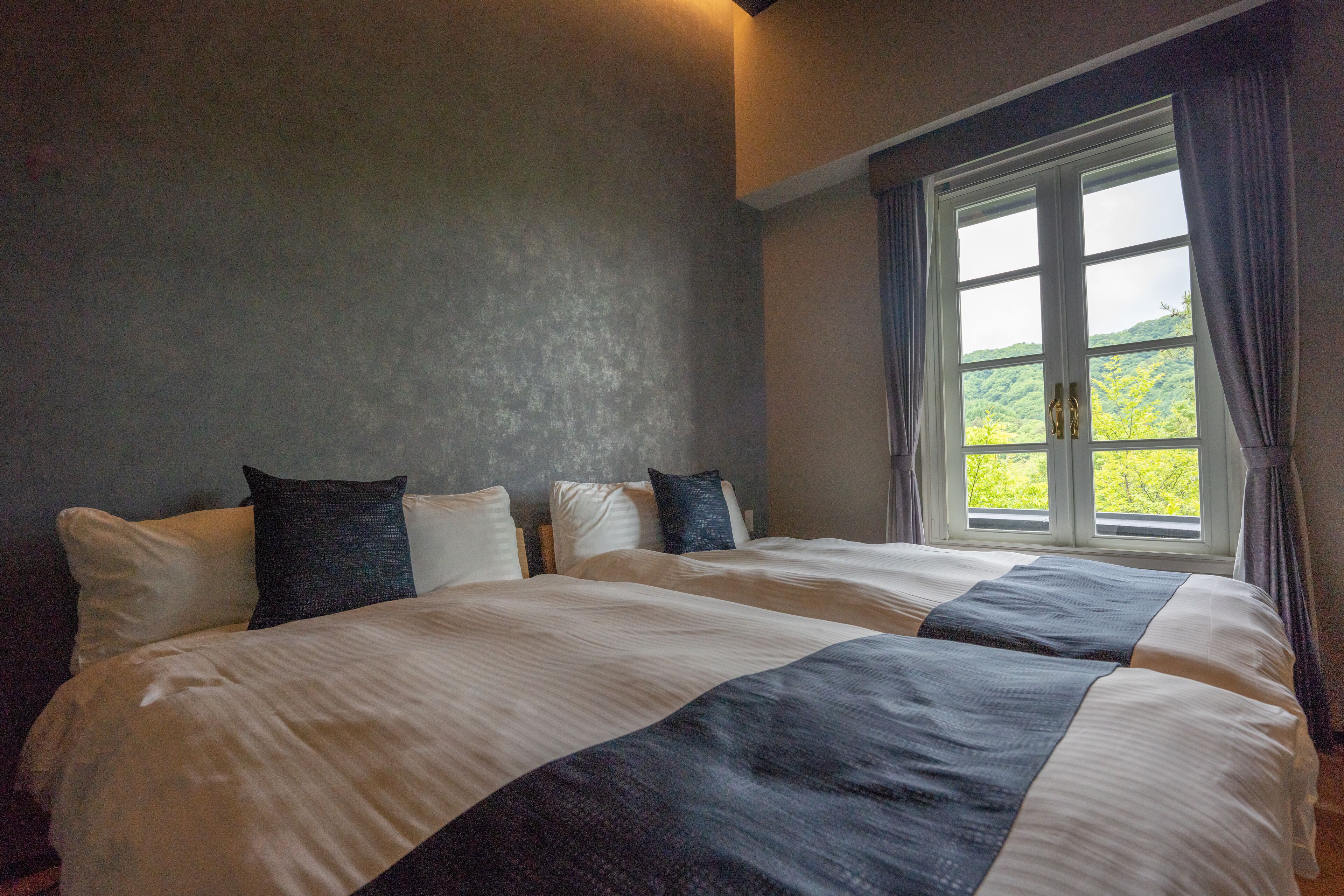 A double bed with white and dark blue bedding, a window with curtains, and a view of greenery outside.